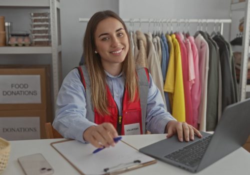 Young woman volunteer using laptop at charity donations center filled with clothes emphasizing community work and caring.