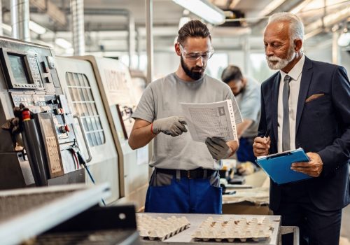Manual worker and senior businessman cooperating while going through paperwork in factory plant.